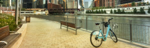 Shared bike parked along a riverwalk with city skyline in the background.