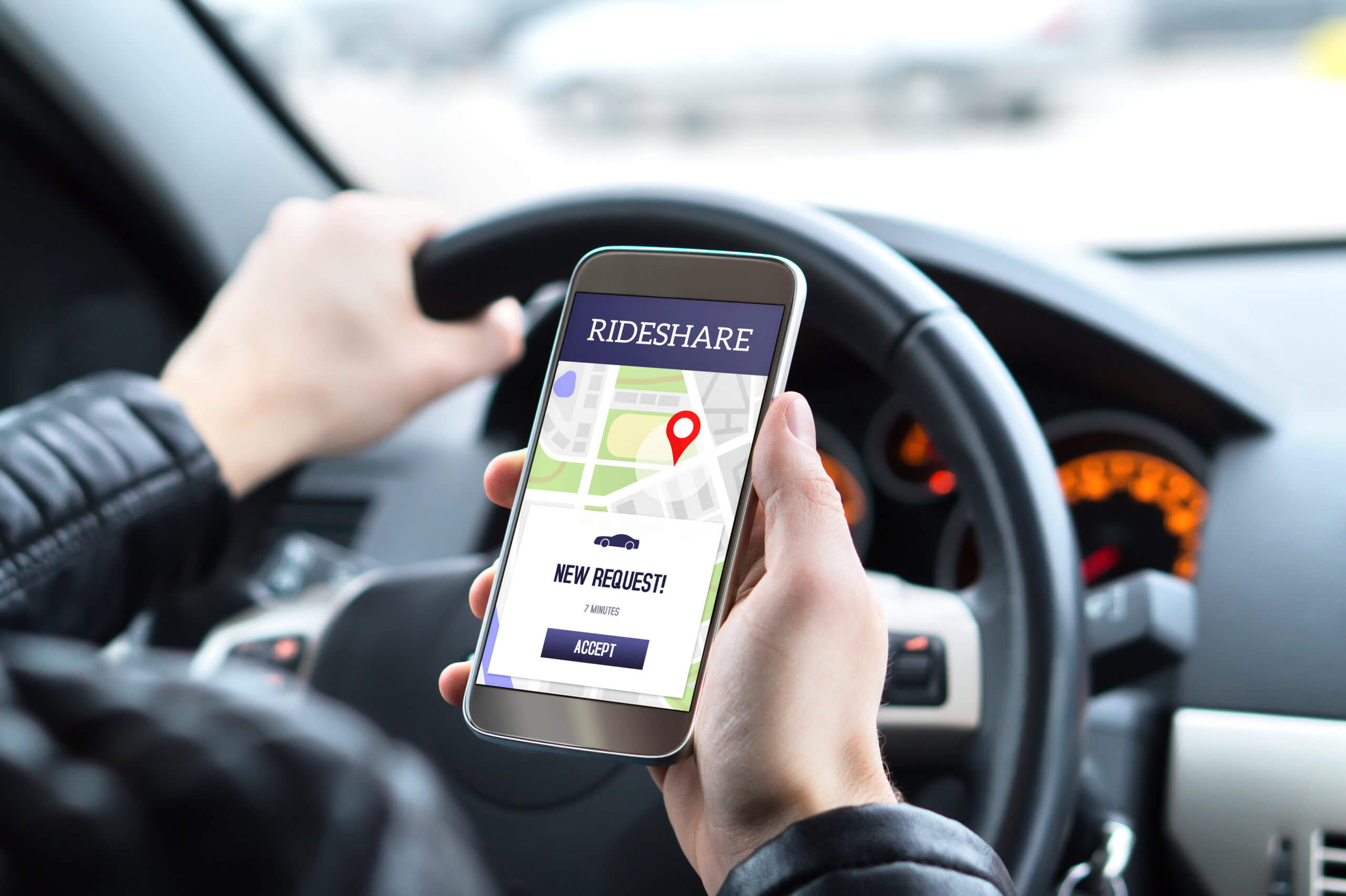 A rideshare driver holding a smartphone with a rideshare app open, showing a new ride request, while sitting behind the wheel of a car.